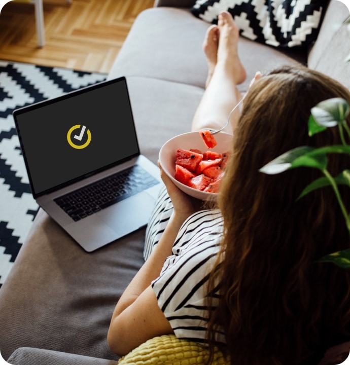 Kobieta leżąca na kanapie, jedząca miskę owoców, obok laptop z logo Norton